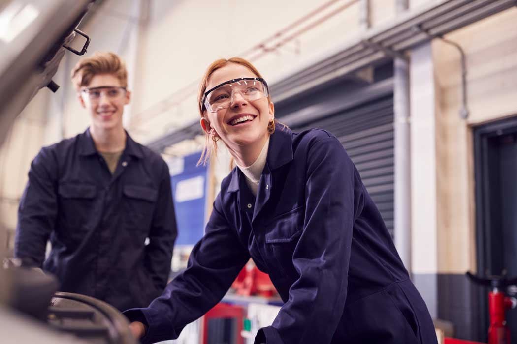 Apprentice mechanic preparing to work on car engine wearing safety glasses looking up and smiling with second apprentice stood by